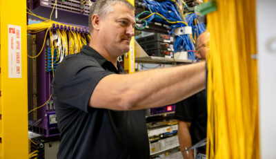 Technician in a dark polo connecting yellow Ethernet cables inside a server rack work cabinet.
