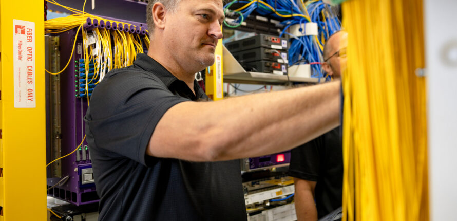 Technician in a dark polo connecting yellow Ethernet cables inside a server rack work cabinet.