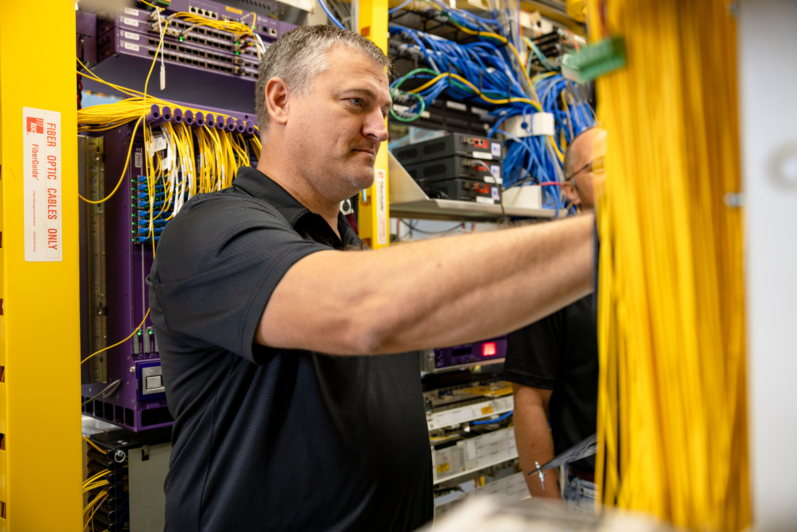 Technician in a dark polo connecting yellow Ethernet cables inside a server rack work cabinet.